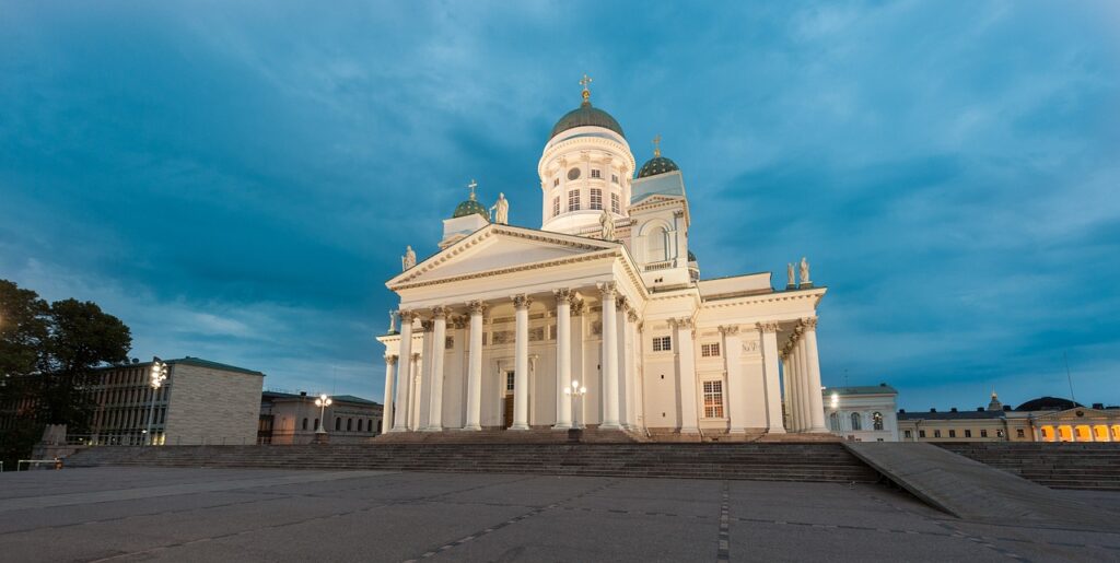 Bilden visar Helsingfors domkyrka. Den har en vitrysk fasad, gröna kupoler och breda trappor. Arkitekturen kännetecknas av en klassicistisk stil och återspeglar de skandinaviska kyrkornas enkelhet.