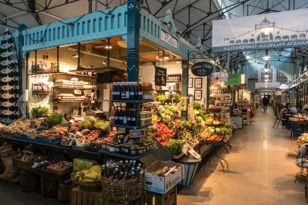 Kuvassa on halli, jossa on erilaisia myyntikojuja. Tarjolla on esimerkiksi hedelmiä, vihanneksia, mehuja ja kalaa.