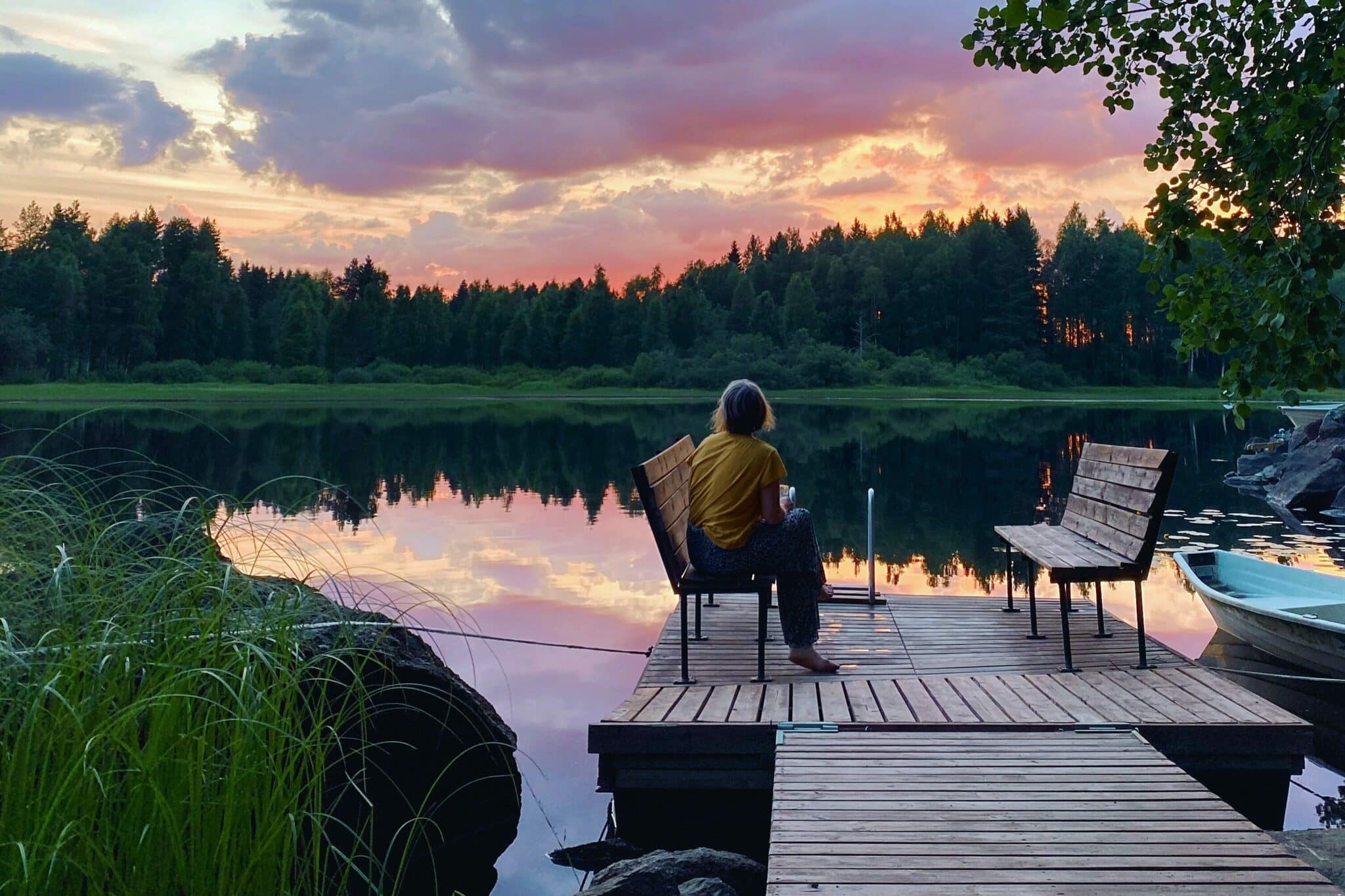 The picture shows a woman, she is sitting on a bench on a jetty. In the background you can see a lake and above it a pink sunset.