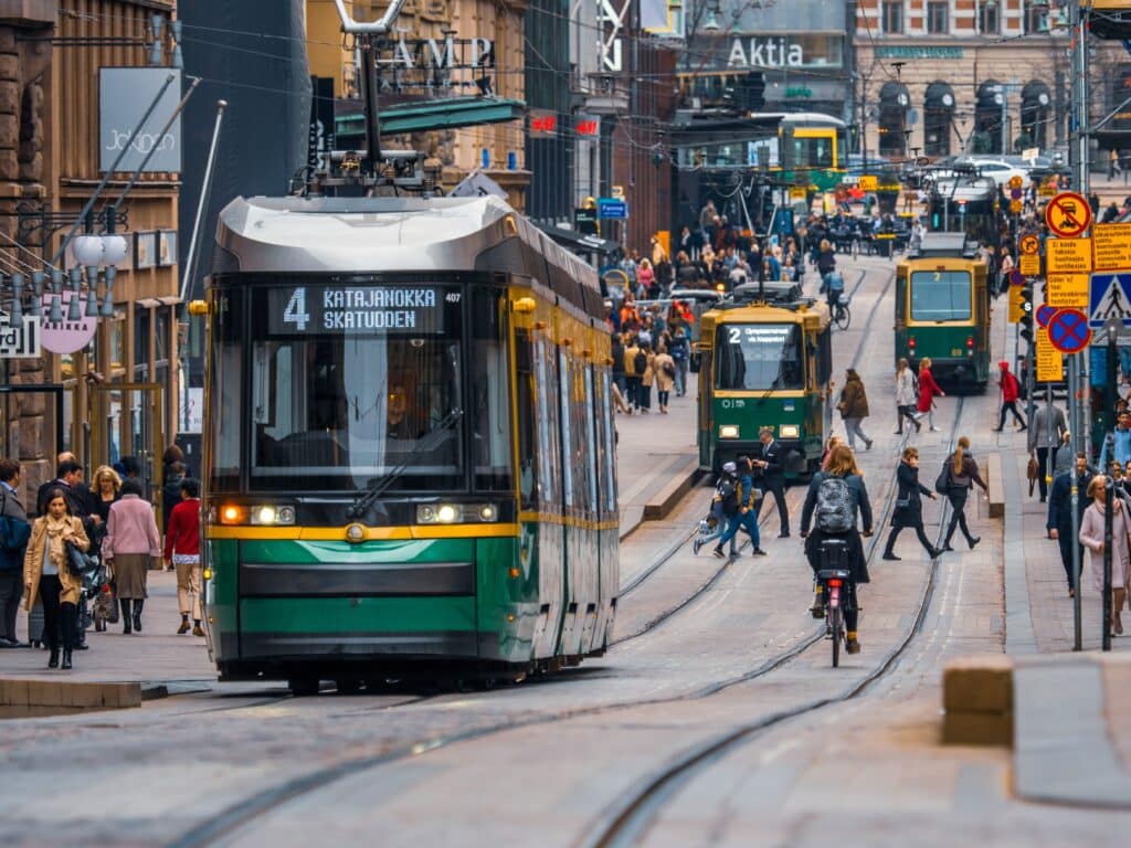 Bilden visar Helsingfors centrum. Du kan se en livlig gata med många människor och flera spårvagnar.